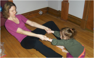 A woman and a toddler sit on a wooden floor facing one another, the woman's legs bent. They are holding hands and rocking back and forth.