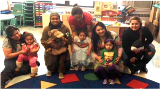 A group of parents and children on a rug with geometrical shapes, smiling and looking at the camera. The children are wearing Halloween costumes.