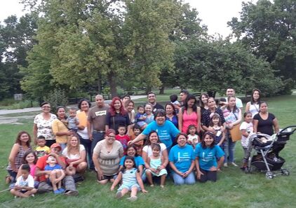 Approximately 40 Latine and Indigenous parents and children stand smiling for a group picture in a park, with leafy trees in the background.