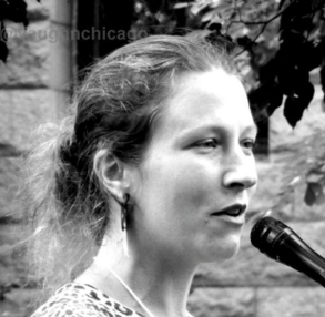 Black and white photo of Kate Duva, a white woman with dark blonde hair in a messy bun and a leopard print sweater, speaking into a microphone.