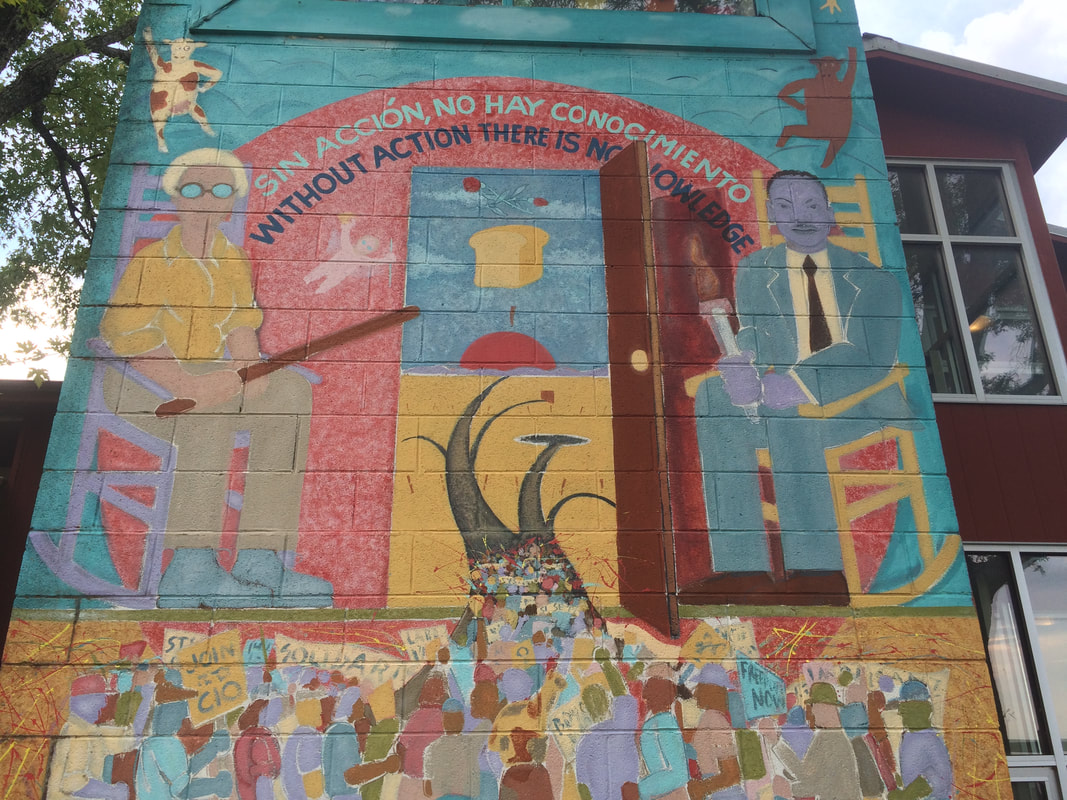 A colorful mural with muted reds, turquoise, and ochre yellow outside on a cinderblock wall depicts Rosa Parks and MLK, Jr in rocking chairs on either side of an open door leading to bread and roses, with protestors underneath. Picture