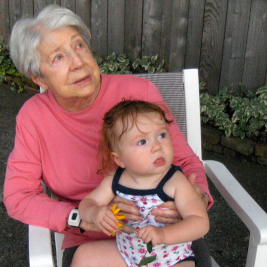 A 90-year-old woman holds on her lap a 9-month old baby wearing a summer onesie and gripping a yellow flower. Both of them are gazing up, looking at something outside the camera's range.
