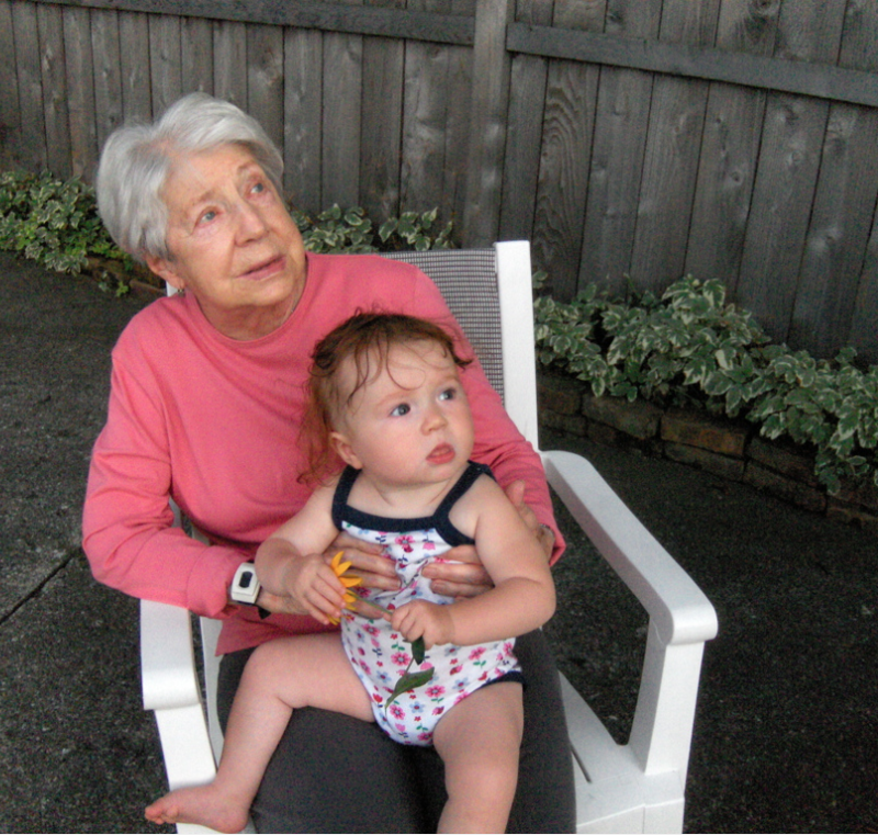 A 90-year-old woman holds on her lap a 9-month old baby wearing a summer onesie and gripping a yellow flower. Both of them are gazing up, looking at something outside the camera's range.